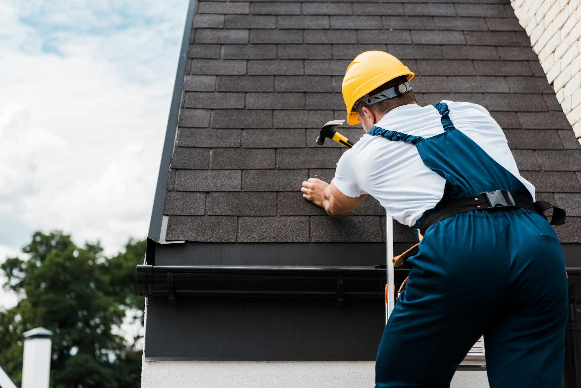 back view of handyman in uniform and helmet repairing roof while standing on ladder